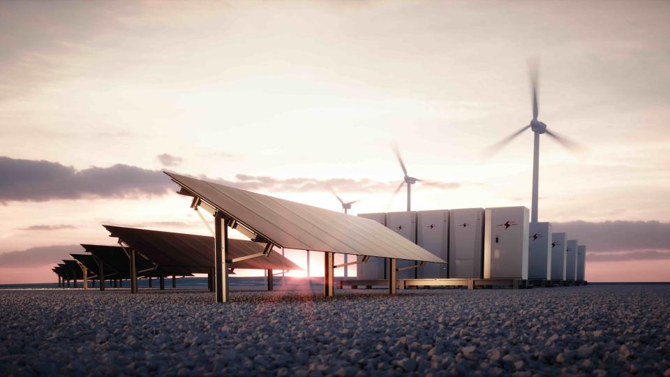 Ground‑mounted solar panel array in the foreground with wind turbines and large battery storage units visible in the background under a cloudy sky.