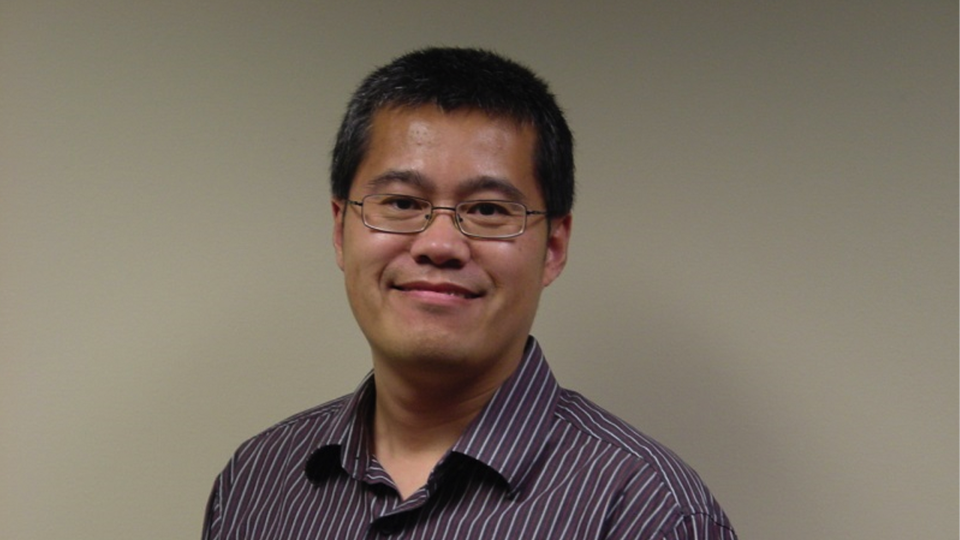 Portrait of a smiling man with short dark hair and glasses wearing a striped button-down shirt against a plain beige background.