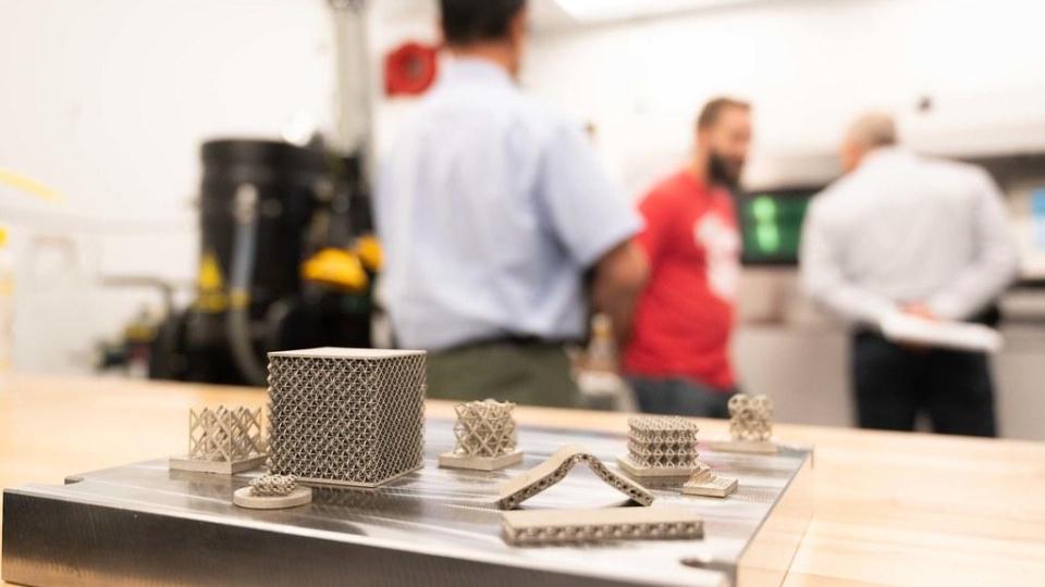 Small metal lattice and cylindrical components arranged on a flat surface in the foreground, with several people standing and talking in a laboratory or workshop space in the background.