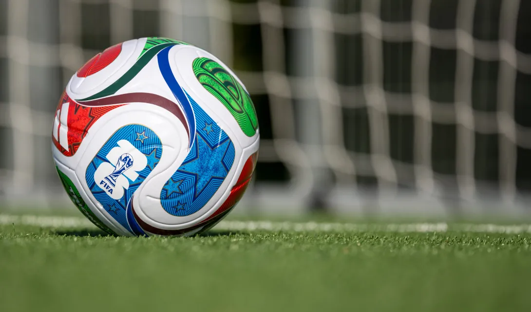 Close-up of a colorful FIFA soccer ball resting on a grass field with a blurred goal net in the background. The ball features blue, green, red, and white geometric designs and the FIFA logo.