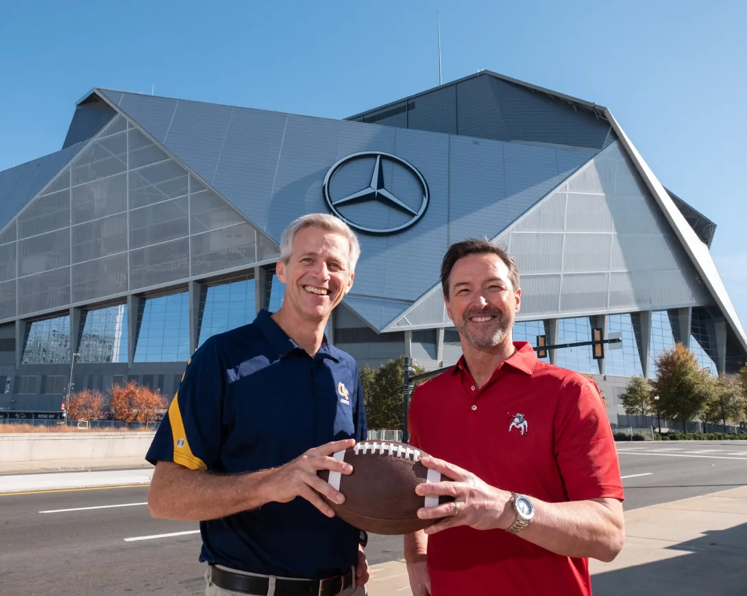 A tall white man wearing a blue GT-branded polo standing next to a slightly shorter man wearing a UGA-branded red polo. They're smiling and both holding a football.