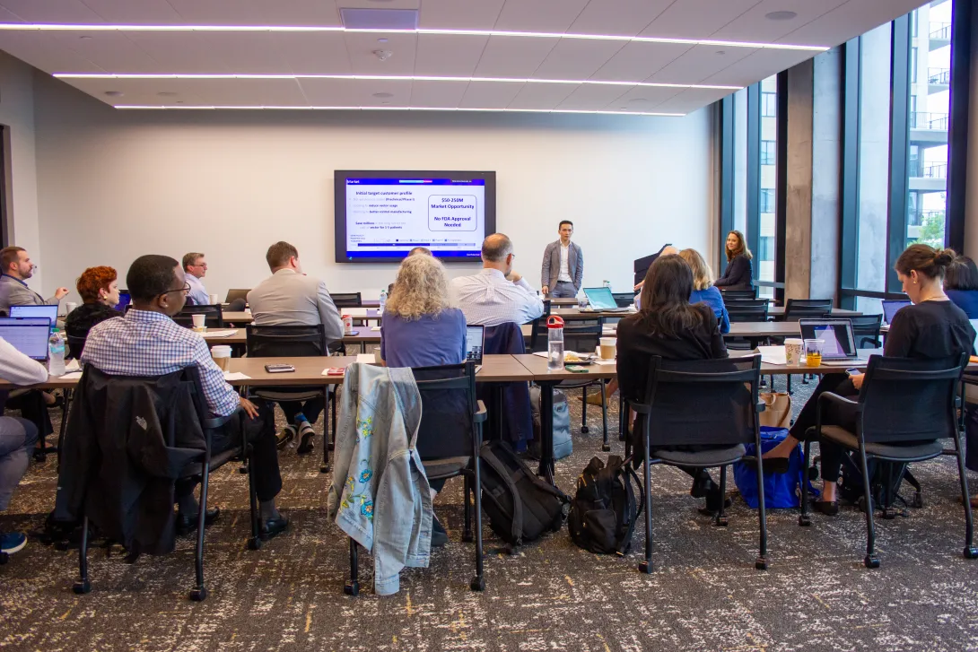 A photo shot from the back of a conference room with people sitting at conference tables while a person at the front of the room shows a presentation on a flat TV screen
