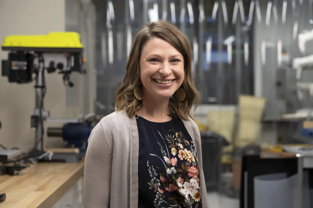 Woman in a workshop environment with industrial equipment and tools in the background, wearing a floral-patterned blouse and light knit cardigan, representing a modern manufacturing or maker space setting.