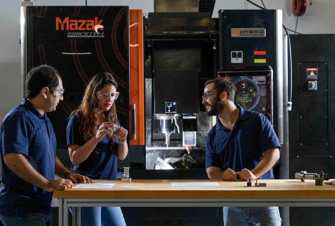 Three individuals standing at a workbench in front of a large Mazak hybrid manufacturing machine, discussing components and technical drawings.