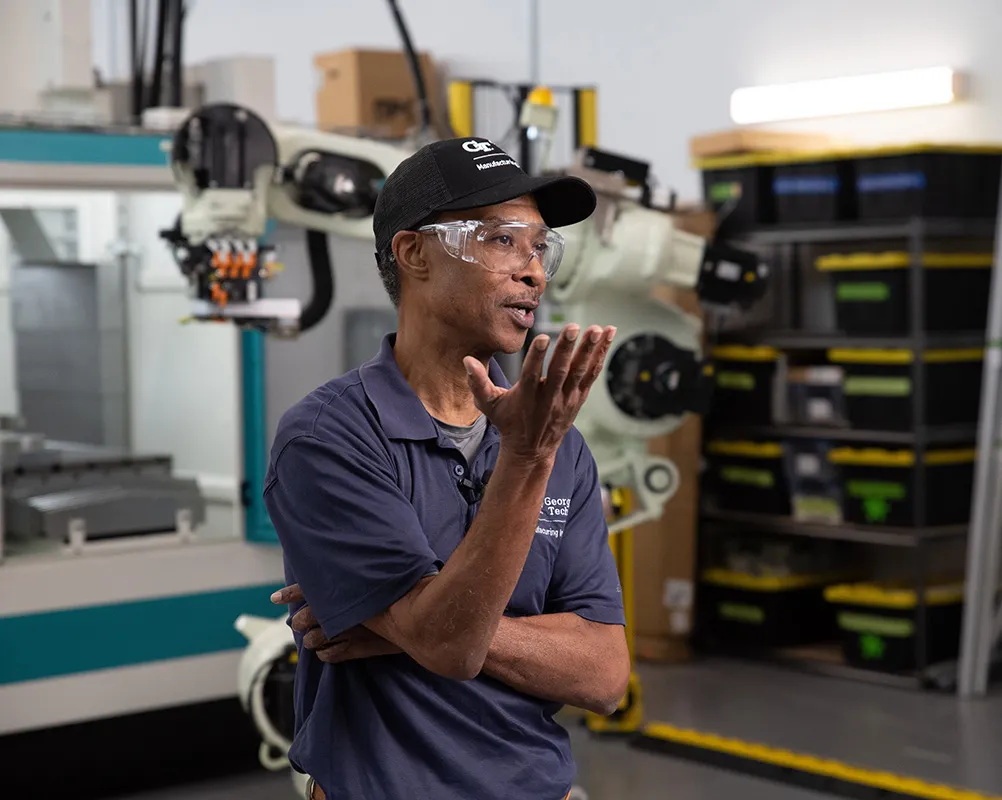A man wearing a cap, safety goggles, and a navy shirt speaks in an industrial setting.