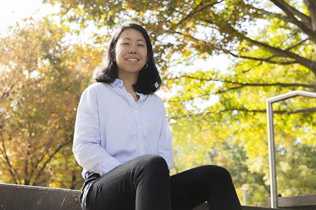 Person sitting outdoors on stone steps wearing a light blue button-up shirt and black pants, with autumn trees and sunlight in the background.