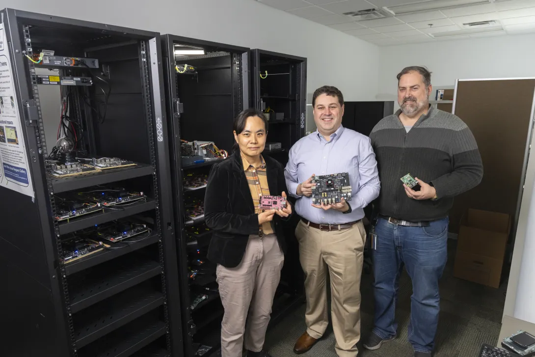 Three researchers stand in front of a rack of computing equipment.