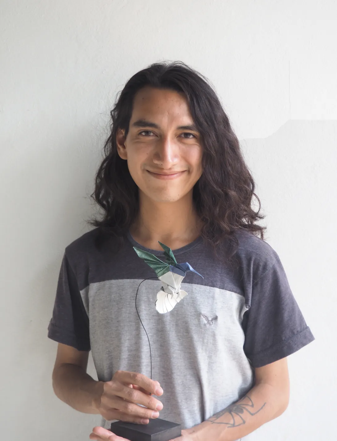 A man with shoulder-length dark hair, and wearing a multicolor t-shirt smiles at the camera while holding an elaborate origami hummingbird. 