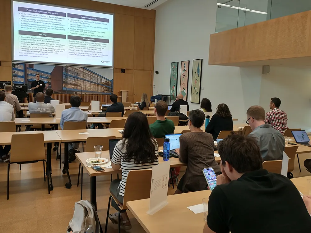 View of a man presenting to a crowd of onlookers in a large meeting room.