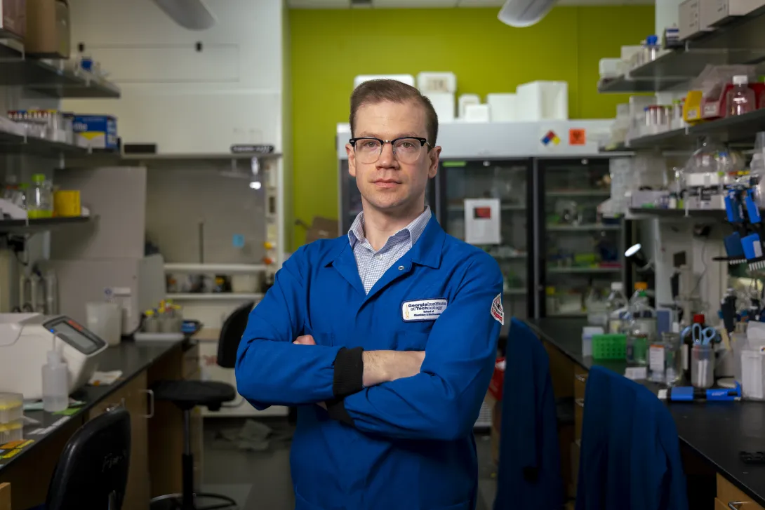 A person wearing a blue lab coat stands with arms crossed in a laboratory filled with shelves of scientific equipment, supplies, and a refrigerator unit in the background.