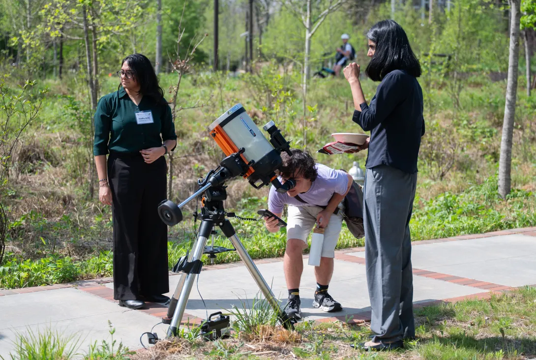 Three people stand outdoors with one person looking at the sun through a telescope
