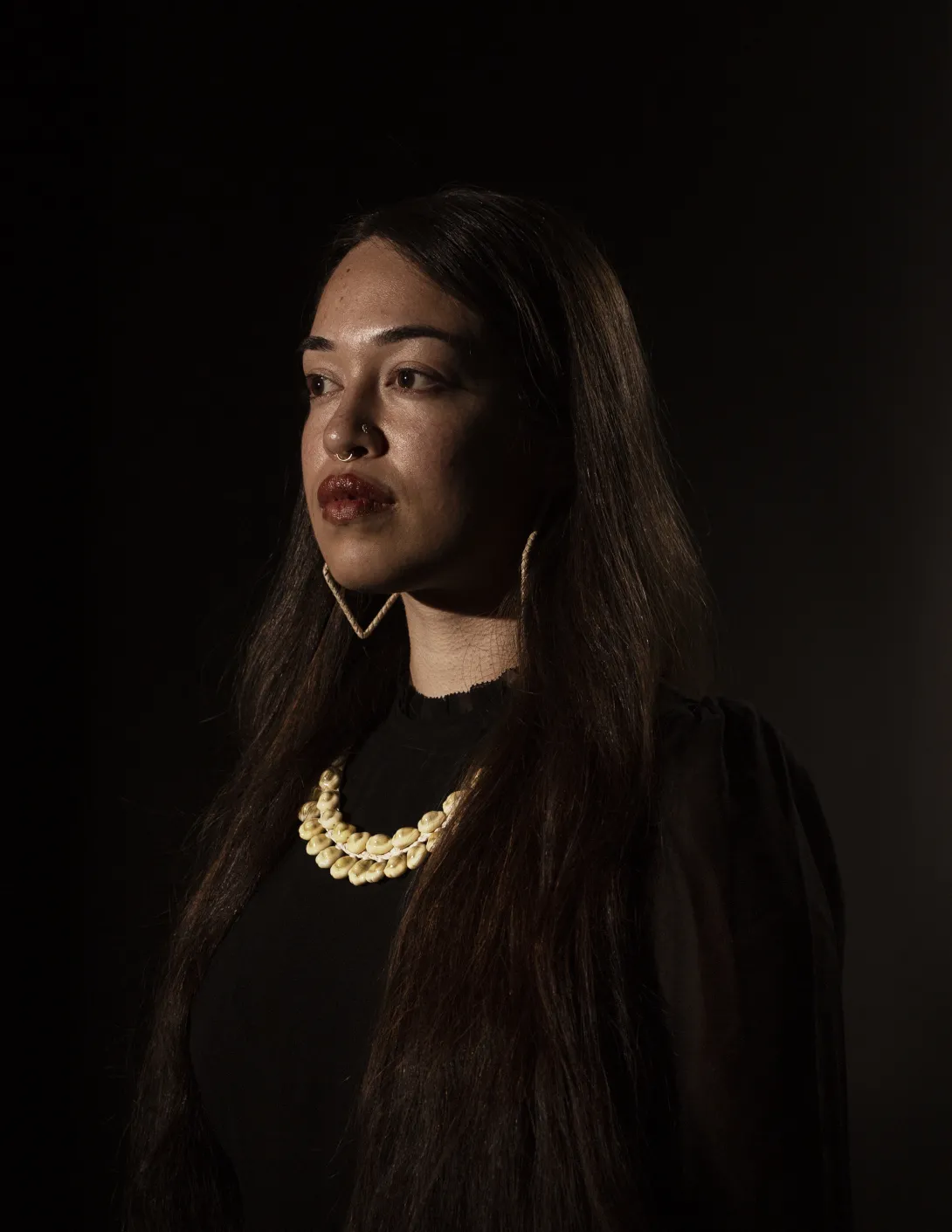 Three-quarter view of woman with long, dark hair looking out of frame. She is wearing large metal earrings and a white, shell necklace.