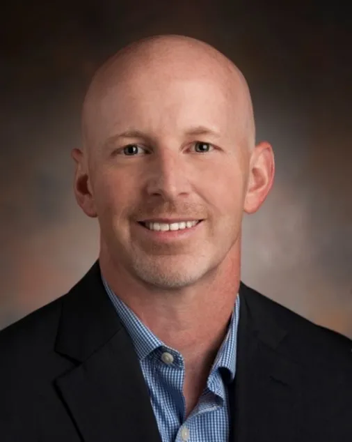 A headshot of a man in a blue shirt and dark blazer