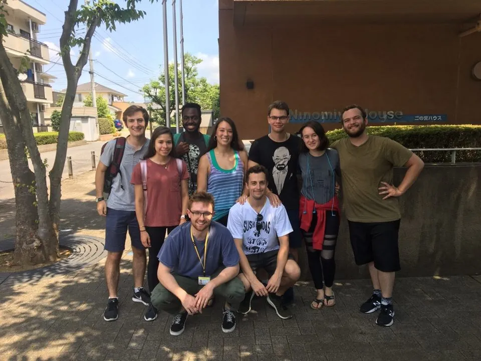 Nine young adults pose together outside a building on a sunny day. Some stand while two kneel in front. They are dressed casually, smiling at the camera, with trees, a sidewalk, and a building entrance sign visible behind them.