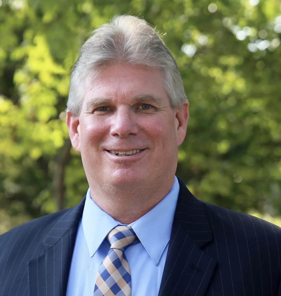 A man wearing a dark blue blazer smiles with greenery in the background.
