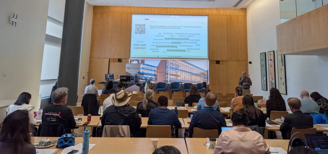 A view inside the Scholars Event Theater of a session of the Sustainability Showcase. A man speaks to a crowd while presenting slides on a large projection screen.