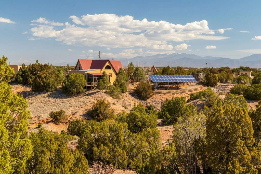A rural residence with solar panels installed outdoors, set among desert vegetation with mountains in the distance.