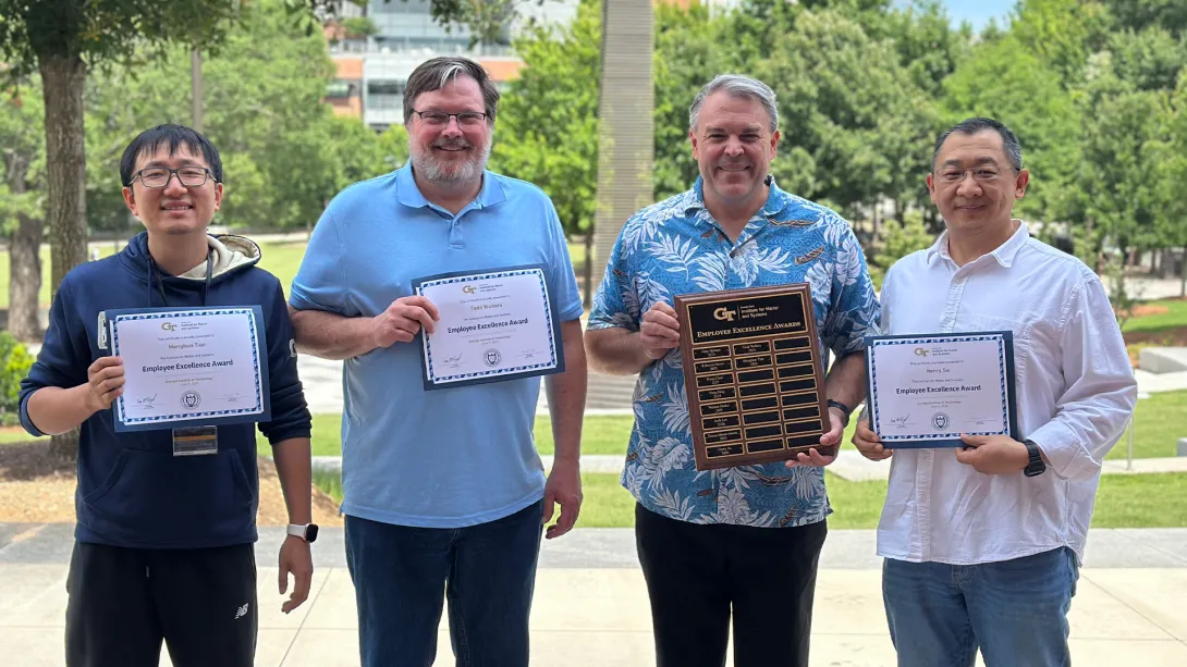 From left to right, Mengkun Tian, Todd Walters, Eric Vogel, and Henry Su stand outdoors holding Employee Excellence Award certificates and a commemorative recognition plaque. The group poses in front of a landscaped campus setting, celebrating their achievements and contributions to the Georgia Tech Institute for Matter and Systems.